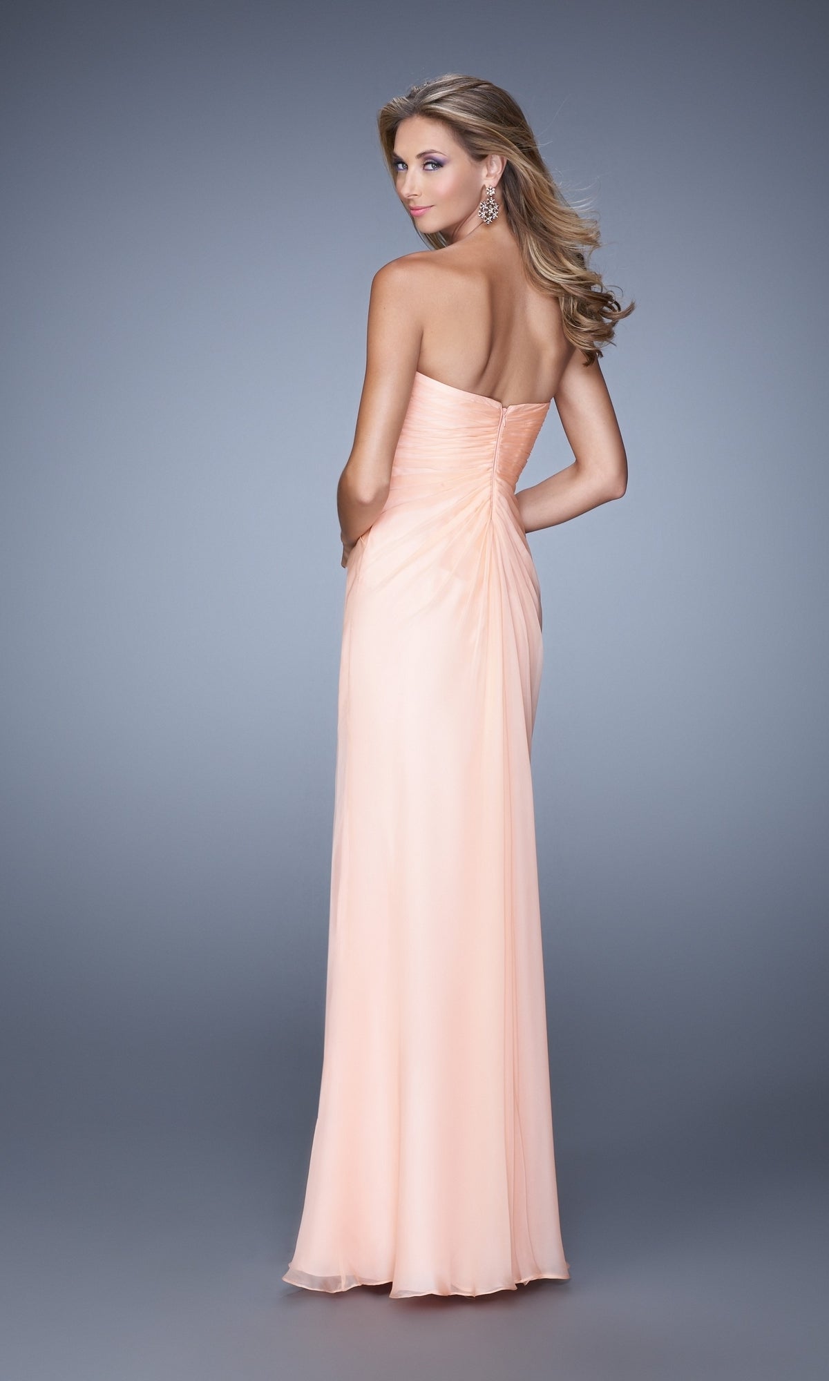 A woman wears the La Femme Dress 21214, a strapless sweetheart peach gown with a ruched bodice and floor-length skirt, shown from the back as she looks over her shoulder against a gray background.